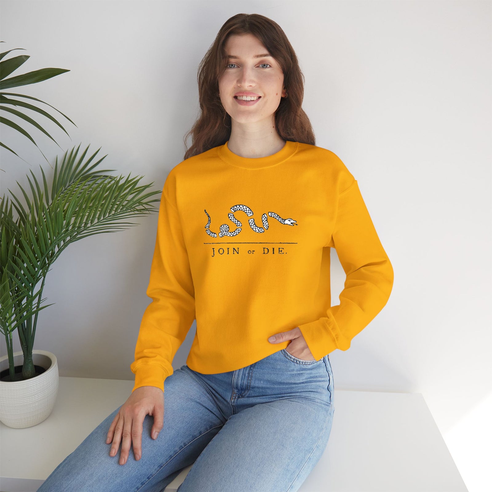 Woman wearing a yellow sweatshirt with a design and text, sitting on a white surface with a plant in the background.