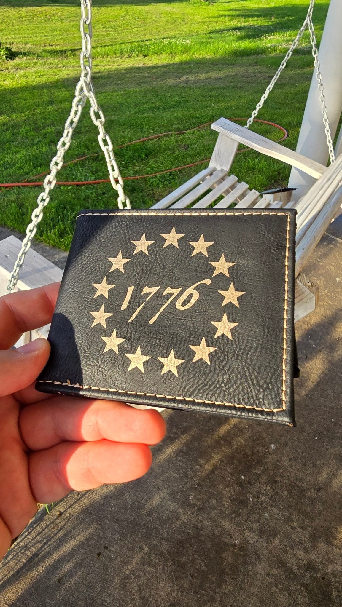 Hand holding the 1776 Betsy Ross black leather wallet in golden evening light on a porch swing, showing engraved 13-star ring and contrast stitching