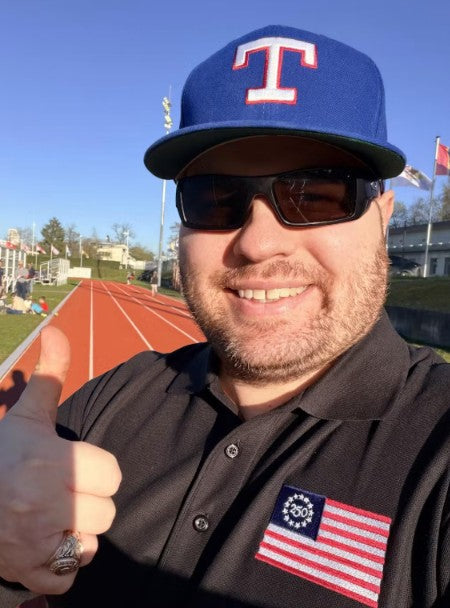 Man wearing a Texas Rangers cap and sunglasses, giving a thumbs-up on a track field.