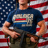 Person wearing a 'America 76' t-shirt holding a rifle in front of an American flag